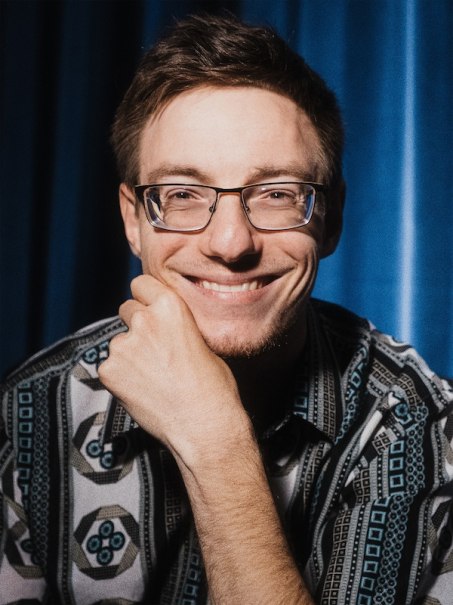 Ein Mann mit Brille lächelt in die Kamera. Er trägt ein gemustertes Hemd und sitzt vor einem blauen Vorhang., © Kulturhaus Schwanen