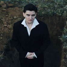 Ein Mann in einem schwarzen Anzug steht in einem offenen Grab und blickt ernst in die Kamera. Die Umgebung ist erdig und grasbewachsen., &copy; links im Bild