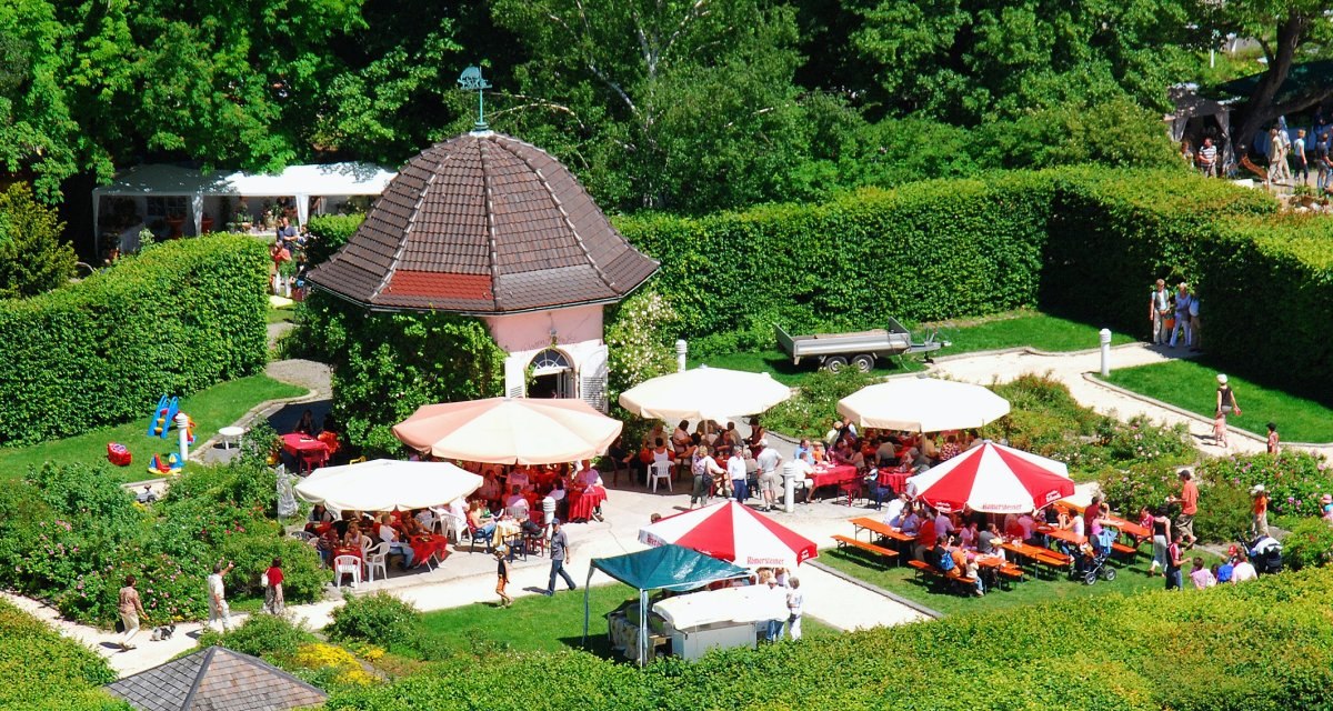 Ein Rosengarten mit Pavillon, umgeben von Hecken. Menschen sitzen unter bunten Sonnenschirmen und genießen das sonnige Wetter., © StaRT GmbH Ein Rosengarten mit Pavillon, umgeben von Hecken. Menschen sitzen unter bunten Sonnenschirmen und genießen das sonnige Wetter., © StaRT GmbH