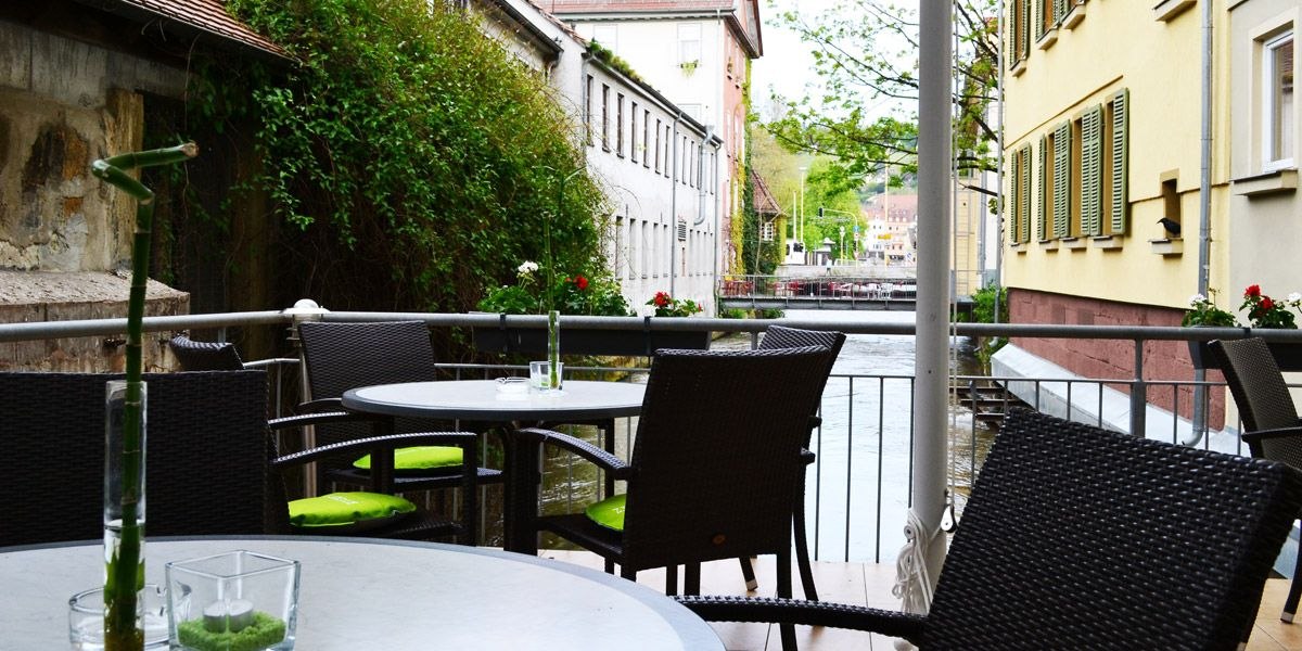 Gem&uuml;tliche Terrasse mit runden Tischen und St&uuml;hlen, Blick auf einen Fluss zwischen zwei Geb&auml;uden, dekoriert mit Pflanzen., &copy; ECOINN Hotel am Campus
