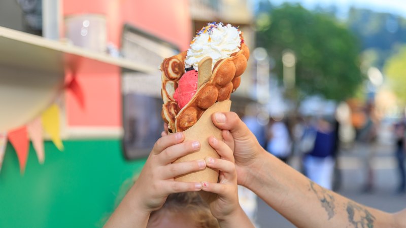 Ein Kind h&auml;lt eine gro&szlig;e Waffel mit Eis und Sahne in den H&auml;nden. Im Hintergrund ist ein Foodtruck und eine belebte Stra&szlig;e zu sehen.