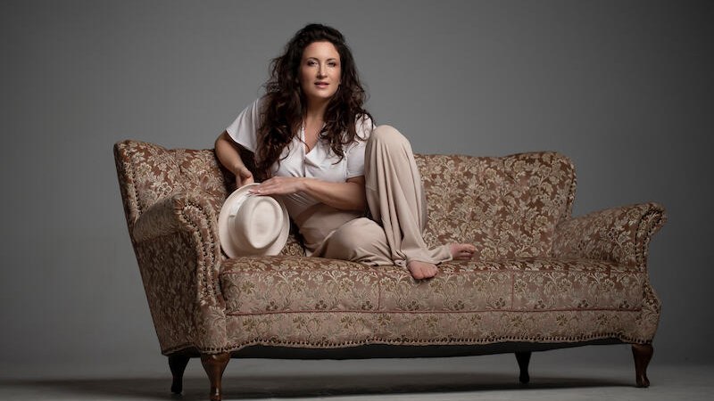 Eine Frau sitzt entspannt auf einem antiken Sofa, h&auml;lt einen Hut in der Hand und tr&auml;gt legere Kleidung. Der Hintergrund ist neutral gehalten., &copy; Sch&ouml;nblick gemeinn&uuml;tzige GmbH