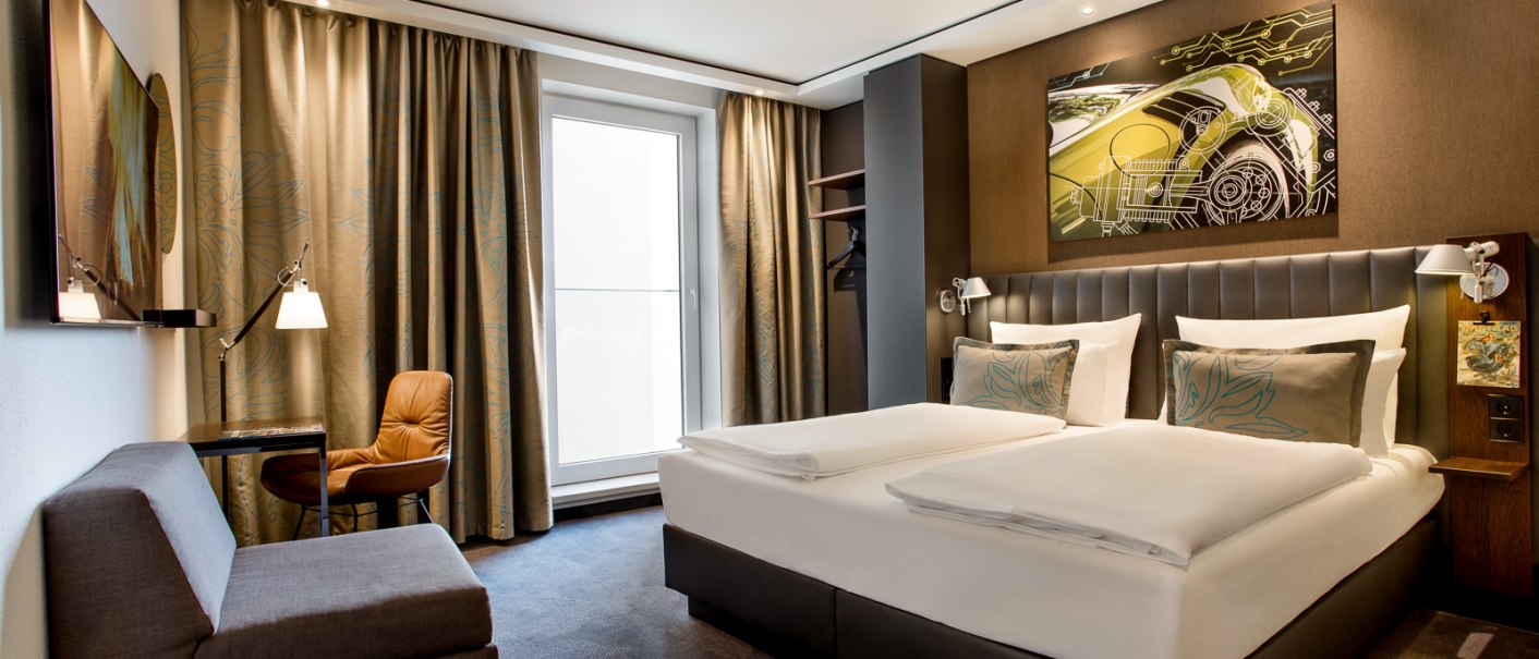 Modernes Hotelzimmer mit Doppelbett, Sofa, Schreibtisch und großem Fenster. An der Wand hängt ein Kunstwerk mit einem Auto-Motiv., © TOMAS Modernes Hotelzimmer mit Doppelbett, Sofa, Schreibtisch und großem Fenster. An der Wand hängt ein Kunstwerk mit einem Auto-Motiv., © TOMAS