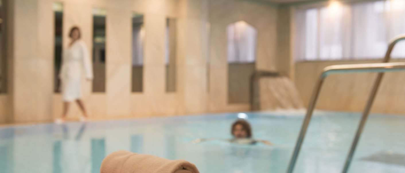 Ein Innenpool mit einem gefalteten Handtuch auf einem roten Tuch im Vordergrund. Im Hintergrund schwimmt eine Person, eine andere geht im Bademantel., © Maritim Hotelgesellschaft mbH