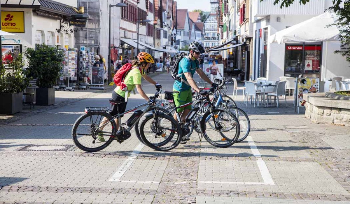 Zwei Radfahrer schieben ihre Fahrräder durch eine belebte Fußgängerzone in Kirchheim unter Teck. Im Hintergrund sind Geschäfte und Cafés zu sehen., © Schwäbische Alb Tourismusverband e.V.
