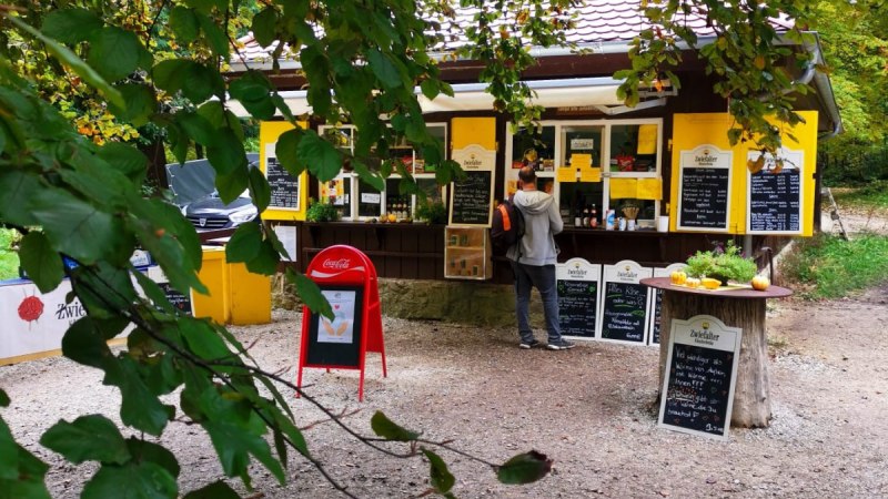 Ein gelber Kiosk im Wald, umgeben von gr&uuml;nen Bl&auml;ttern. Ein Kunde steht davor. Auf Tafeln sind Angebote geschrieben.
