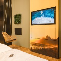 Modernes Hotelzimmer mit Hängesessel, Fernseher mit Strandbild, kleiner Küchenzeile und dekorativem Pflanzenbild an der Wand., © TOMAS Modernes Hotelzimmer mit Hängesessel, Fernseher mit Strandbild, kleiner Küchenzeile und dekorativem Pflanzenbild an der Wand., © TOMAS