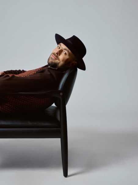 Eine Person mit Hut lehnt entspannt auf einem schwarzen Stuhl, seitlich liegend, vor einem grauen Hintergrund., © Scala Kultur Live gGmbH