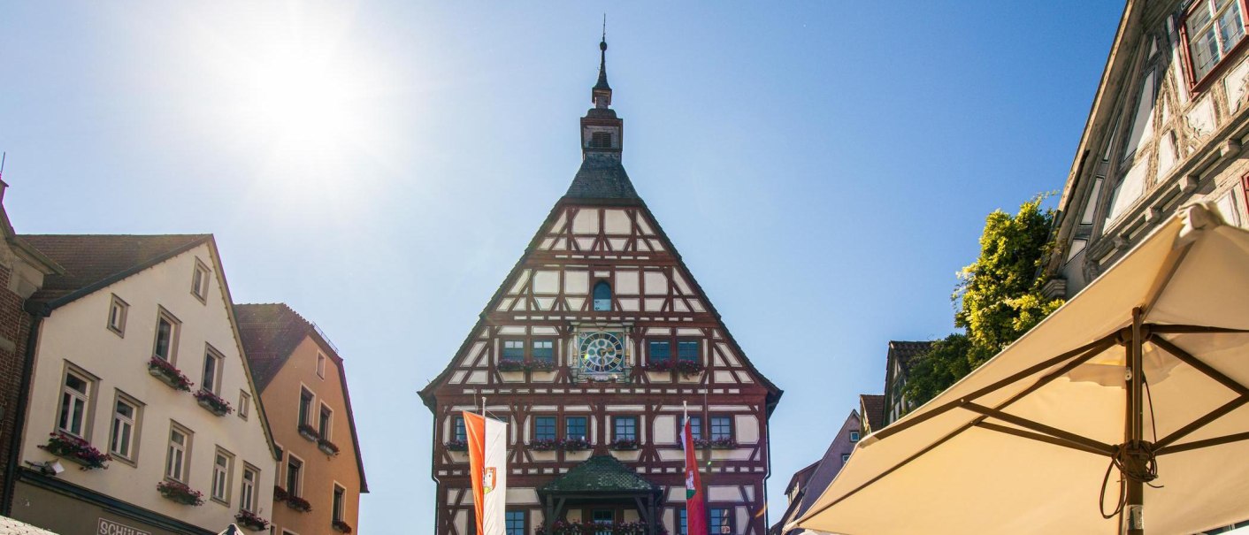 Das Fachwerk-Rathaus von Besigheim steht im Sonnenschein auf dem Marktplatz, umgeben von historischen Geb&auml;uden und einem Sonnenschirm., &copy; Stuttgart-Marketing GmbH, Sarah Schmid