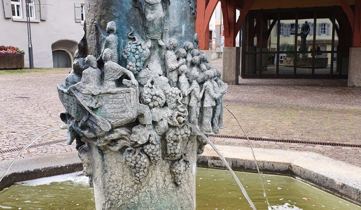 Brunnen mit detaillierten Skulpturen und Wasserspeiern, umgeben von Fachwerkhäusern und einem gepflasterten Platz., © Kulturamt Stadt Plochingen Brunnen mit detaillierten Skulpturen und Wasserspeiern, umgeben von Fachwerkhäusern und einem gepflasterten Platz., © Kulturamt Stadt Plochingen