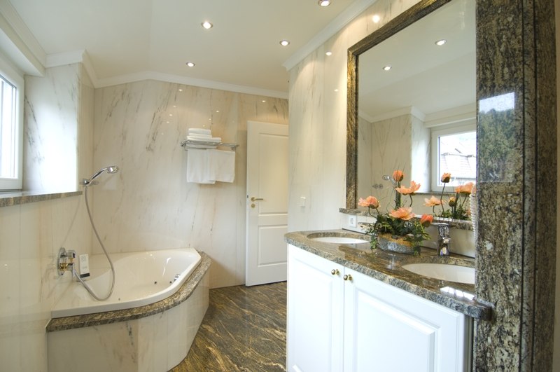 Elegantes Badezimmer mit Whirlpool, Doppelwaschbecken, Marmorwänden und Blumenarrangement. Helle Beleuchtung und Fenster sorgen für eine angenehme Atmosphäre., © Jeanette Bender Elegantes Badezimmer mit Whirlpool, Doppelwaschbecken, Marmorwänden und Blumenarrangement. Helle Beleuchtung und Fenster sorgen für eine angenehme Atmosphäre., © Jeanette Bender