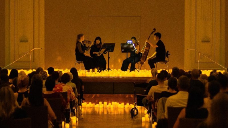 Ein Streichquartett spielt auf einer kerzenbeleuchteten Bühne vor einem sitzenden Publikum in einem Konzertsaal., © FEVER Ein Streichquartett spielt auf einer kerzenbeleuchteten Bühne vor einem sitzenden Publikum in einem Konzertsaal., © FEVER