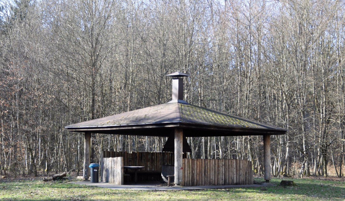 Eine h&ouml;lzerne Grillh&uuml;tte mit einem Dach und einem Schornstein steht auf einer Lichtung, umgeben von kahlen B&auml;umen., &copy; Natur.Nah. Sch&ouml;nbuch & Heckeng&auml;u