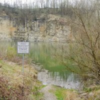 Ein kleiner See mit steilen Felswänden, umgeben von Bäumen. Ein Warnschild weist auf Gefahren hin. Der Himmel ist bewölkt., © Hans Sukowski Ein kleiner See mit steilen Felswänden, umgeben von Bäumen. Ein Warnschild weist auf Gefahren hin. Der Himmel ist bewölkt., © Hans Sukowski