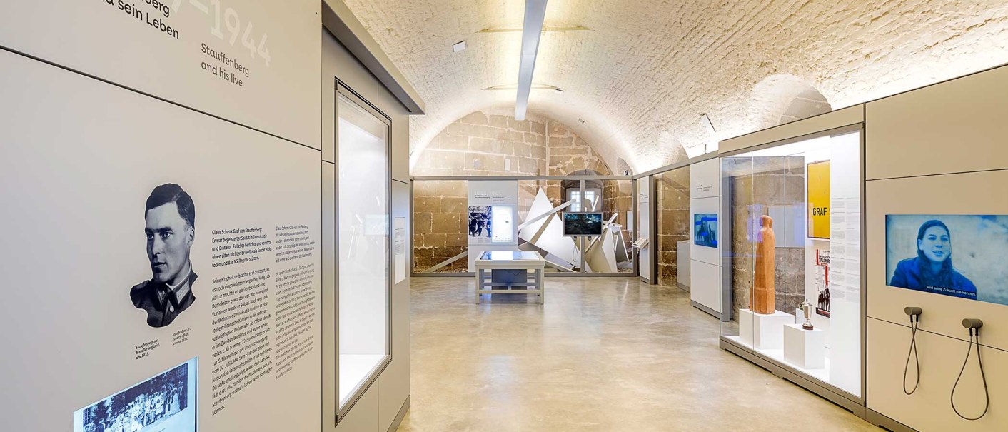 Innenansicht der Stauffenberg-Erinnerungsstätte mit modernen Ausstellungsstücken, Informationstafeln und Bildschirmen in einem gewölbten Raum., © Haus der Geschichte Baden-Württemberg Innenansicht der Stauffenberg-Erinnerungsstätte mit modernen Ausstellungsstücken, Informationstafeln und Bildschirmen in einem gewölbten Raum., © Haus der Geschichte Baden-Württemberg