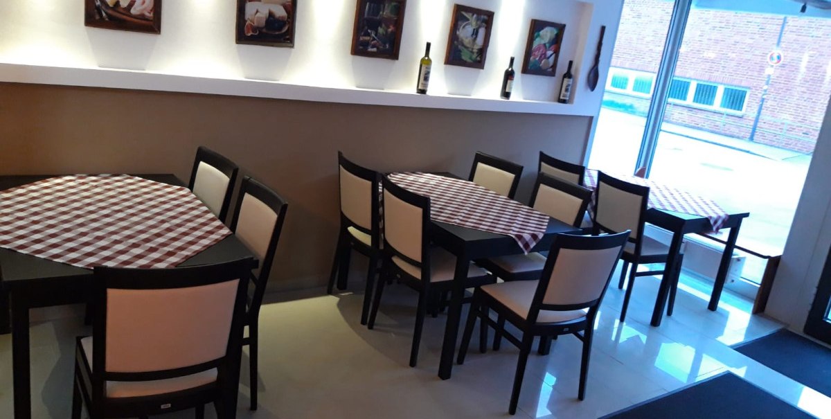 Restaurant mit karierten Tischdecken, schwarzen Stühlen und Wandbildern. Helle Beleuchtung und große Fensterfront.
