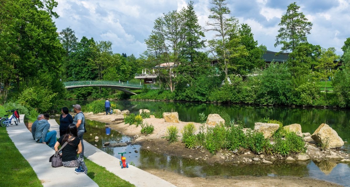 Menschen entspannen an einem Flussufer in einem gr&uuml;nen Park. Eine Br&uuml;cke und B&auml;ume sind im Hintergrund zu sehen., &copy; Remstal Tourismus e.V.