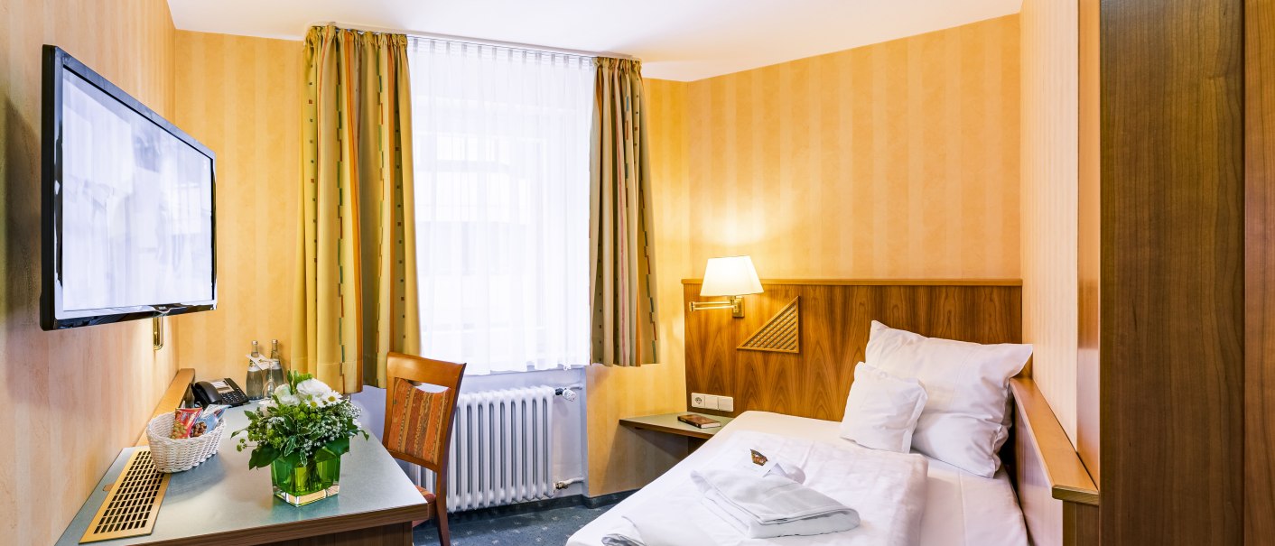 Kleines Hotelzimmer mit Einzelbett, Schreibtisch, Fernseher und Fenster mit Vorhängen. Ein Blumenarrangement steht auf dem Tisch., © TOMAS