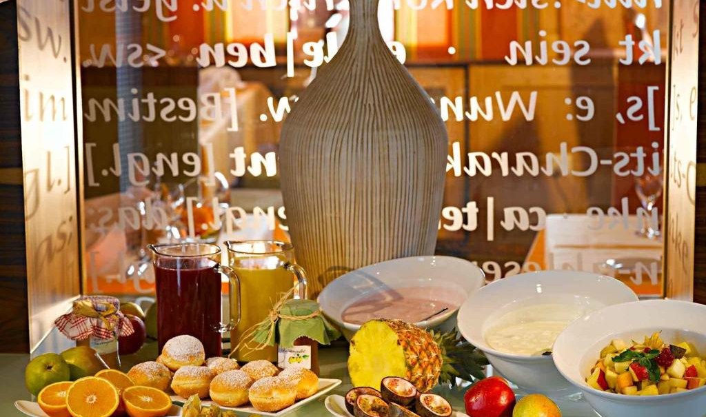 Ein Frühstücksbuffet mit frischem Obst, Gebäck, Joghurt und Säften. Im Hintergrund eine große Vase und dekorative Schrift an der Wand., © Nödinger Gastro GmbH