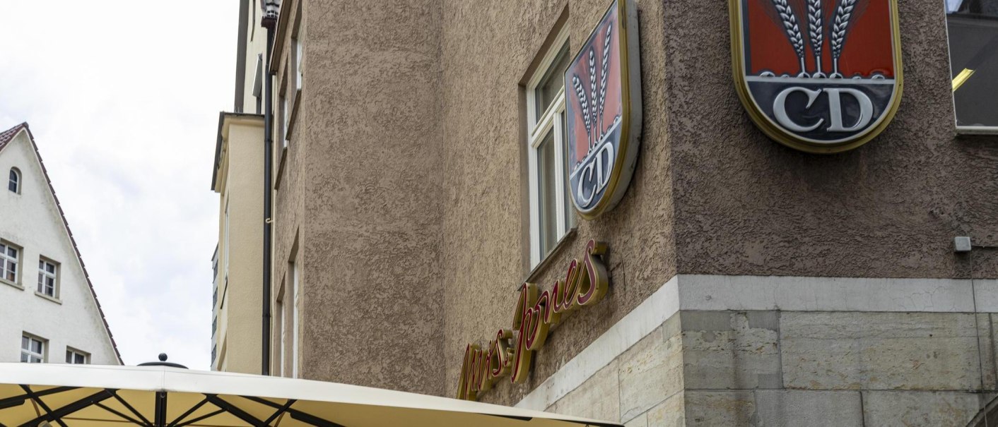 Ein Gebäude mit dem Schriftzug 'Mrs Jones' und einem Logo mit Ähren und den Buchstaben 'CD'. Ein Sonnenschirm ist im Vordergrund sichtbar., © SMG, Sarah Schmid Ein Gebäude mit dem Schriftzug 'Mrs Jones' und einem Logo mit Ähren und den Buchstaben 'CD'. Ein Sonnenschirm ist im Vordergrund sichtbar., © SMG, Sarah Schmid