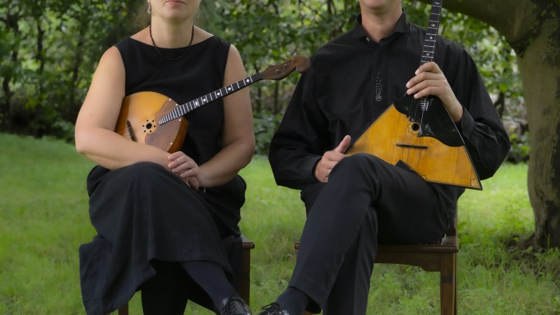 Zwei Musiker mit Balalaika und Domra sitzen im Freien auf St&uuml;hlen. Sie tragen schwarze Kleidung und sind von gr&uuml;ner Natur umgeben., &copy; Exprompt