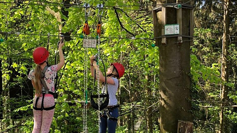 Zwei Personen mit roten Helmen klettern im Skypark Kletterwald in Schwäbisch Gmünd. Sie sind mit Sicherheitsgurten gesichert und von Bäumen umgeben., © SKYPARK – epia-store.de GbR Zwei Personen mit roten Helmen klettern im Skypark Kletterwald in Schwäbisch Gmünd. Sie sind mit Sicherheitsgurten gesichert und von Bäumen umgeben., © SKYPARK – epia-store.de GbR