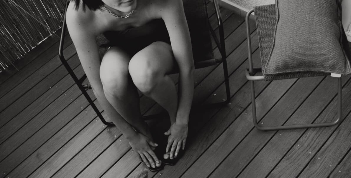 Eine Person sitzt auf einem Balkonstuhl und beugt sich zu einem Hund hinunter. Der Balkon hat Holzboden und eine Bambuswand. Schwarz-weiß-Foto., © Peter Oliver Wolff / DAS EDITH