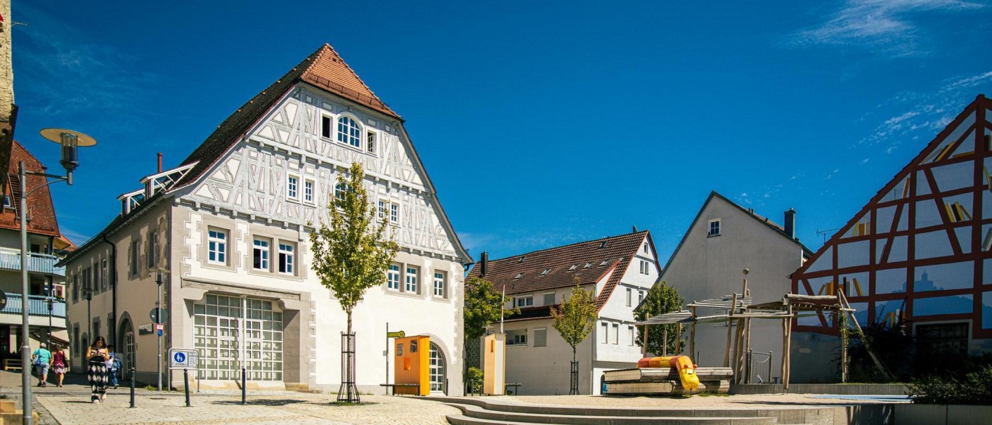 Historisches Fachwerkhaus am Marktplatz von Vaihingen an der Enz, umgeben von modernen Gebäuden und einem kleinen Spielplatz, bei strahlend blauem Himmel., © Stuttgart-Marketing GmbH, Sarah Schmid