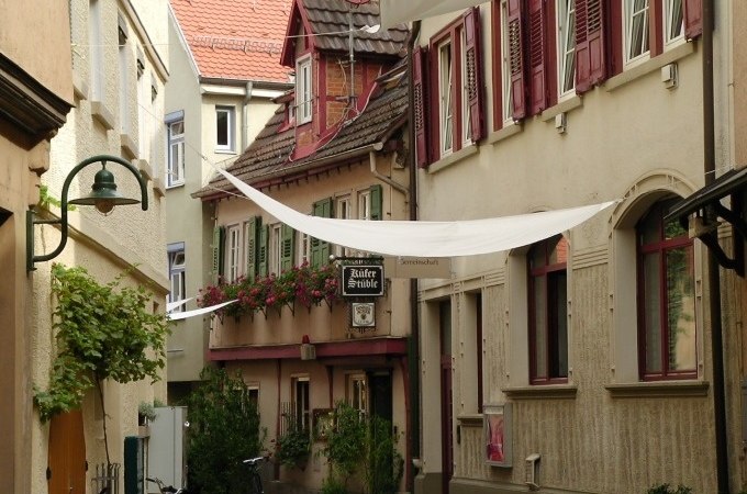 Schmale Gasse mit historischen Gebäuden, Blumen an den Fenstern und weißen Sonnensegeln. Ein Schild weist auf ein Lokal hin., © Cool-Tours StattReisen
