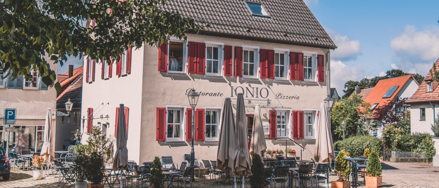 Ein Restaurant mit roten Fensterläden und Außensitzbereich auf einem gepflasterten Platz, umgeben von Bäumen und blauem Himmel., © Stadt Welzheim Ein Restaurant mit roten Fensterläden und Außensitzbereich auf einem gepflasterten Platz, umgeben von Bäumen und blauem Himmel., © Stadt Welzheim