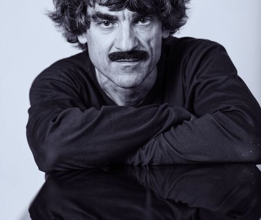 Ein Mann mit lockigem Haar und Schnurrbart lehnt sich auf einen Tisch. Sein Gesicht spiegelt sich auf der glänzenden Oberfläche wider., © Thomas Scheytt Ein Mann mit lockigem Haar und Schnurrbart lehnt sich auf einen Tisch. Sein Gesicht spiegelt sich auf der glänzenden Oberfläche wider., © Thomas Scheytt