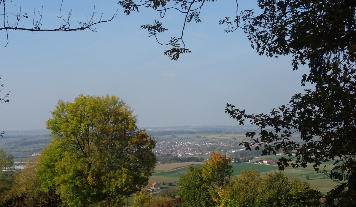 Blick von einem Hügel ins Tal, umrahmt von Bäumen. Im Hintergrund ist ein Dorf zu sehen, eingebettet in eine weite Landschaft unter blauem Himmel., © Foto: Cornelia Steinbach Blick von einem Hügel ins Tal, umrahmt von Bäumen. Im Hintergrund ist ein Dorf zu sehen, eingebettet in eine weite Landschaft unter blauem Himmel., © Foto: Cornelia Steinbach