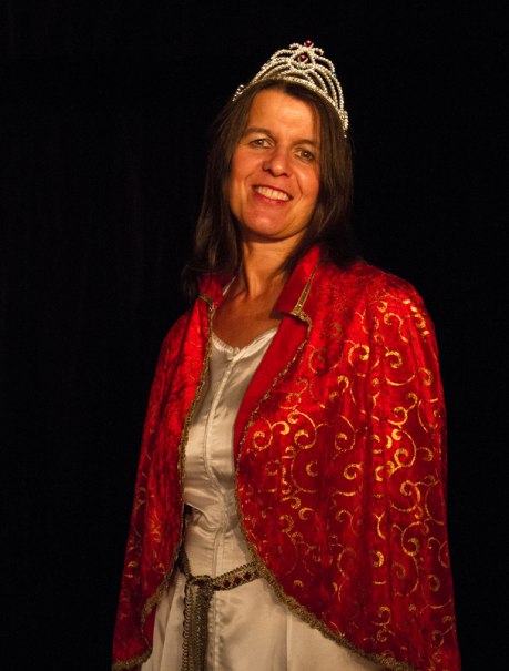 Eine Frau in einem roten, goldverzierten Umhang und einer Krone l&auml;chelt vor einem dunklen Hintergrund., &copy; Galli Theater Backnang