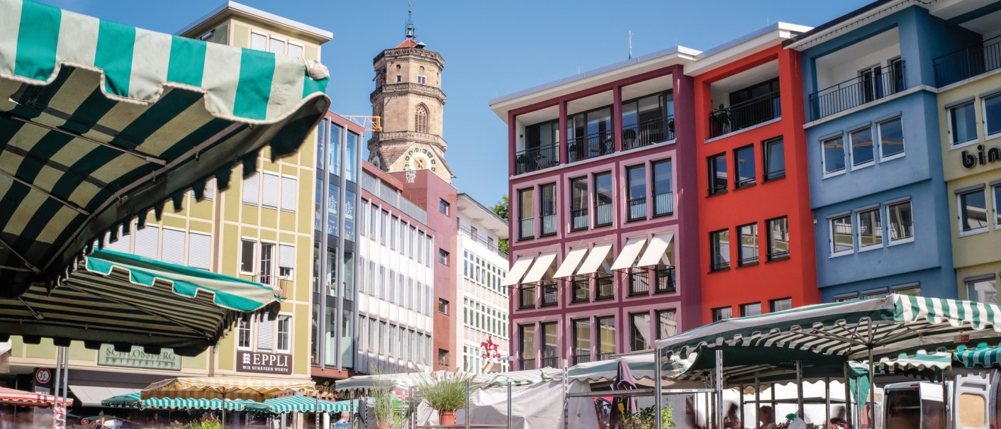 Bunte Gebäude und Marktstände auf einem Marktplatz, mit einem Kirchturm im Hintergrund unter blauem Himmel., © SMG, trickytine
