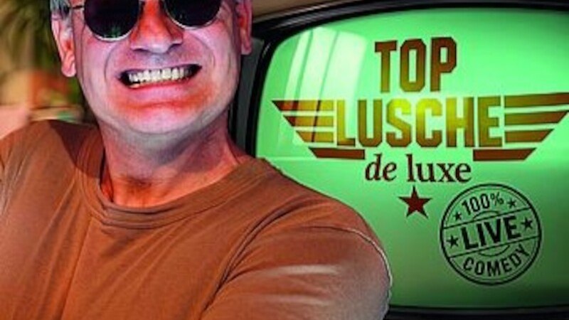 Ein Mann mit roter Mütze und Sonnenbrille lächelt breit vor einem Fernseher mit der Aufschrift 'Top Lusche de luxe'., © Mauerwerk GmbH Ein Mann mit roter Mütze und Sonnenbrille lächelt breit vor einem Fernseher mit der Aufschrift 'Top Lusche de luxe'., © Mauerwerk GmbH