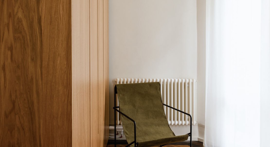 Minimalistischer Raum mit Holzschrank, grünem Stuhl und Parkettboden. Ein Fenster mit weißen Vorhängen lässt Licht herein., © Peter Oliver Wolff / DAS EDITH