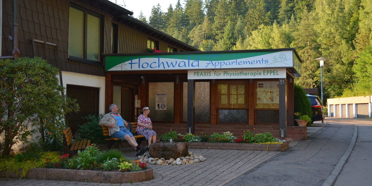 Gebäude mit Schild 'Hochwald Appartements' und 'Praxis für Physiotherapie EPPEL'. Zwei Personen sitzen auf einer Bank, umgeben von Pflanzen und Bäumen., © Hotel Hochwald