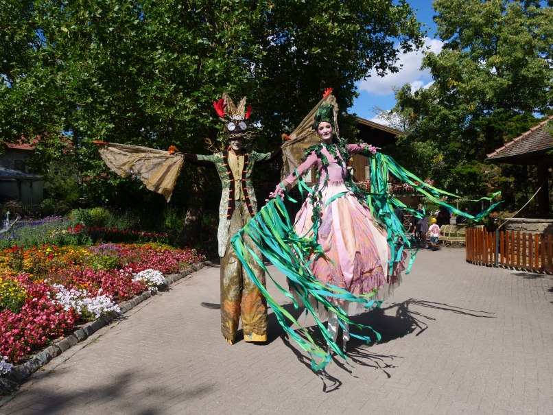 Zwei Personen auf Stelzen in fantasievollen Kost&uuml;men spazieren durch einen bl&uuml;henden Garten. Bunte Blumen und B&auml;ume umgeben den Weg., &copy; Erlebnispark Tripsdrill
