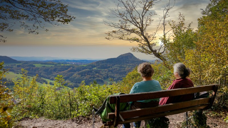 Zwei Personen sitzen auf einer Bank am Aussichtspunkt Luginsland und genießen den Blick auf die hügelige Landschaft im Sonnenuntergang., © Foto: Mario Klaiber
