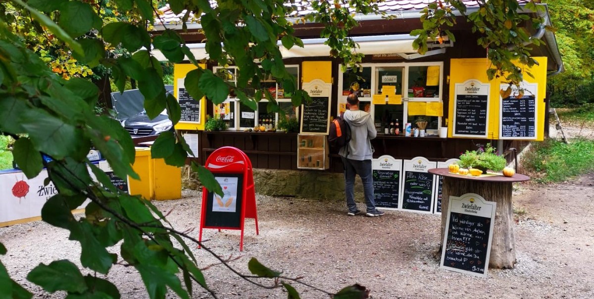 Ein gelber Kiosk im Wald, umgeben von gr&uuml;nen Bl&auml;ttern. Ein Kunde steht davor. Auf Tafeln sind Angebote geschrieben.