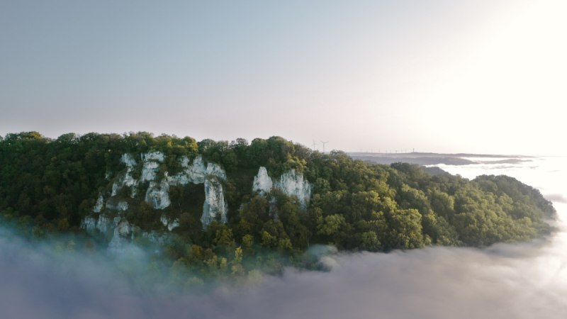 Ein bewaldeter Hügel mit markanten Felsformationen erhebt sich über Nebel. Im Hintergrund sind Windräder zu sehen, die in der Ferne stehen., © Landkreis Göppingen Ein bewaldeter Hügel mit markanten Felsformationen erhebt sich über Nebel. Im Hintergrund sind Windräder zu sehen, die in der Ferne stehen., © Landkreis Göppingen