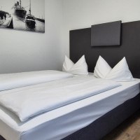 Modernes Hotelzimmer mit Doppelbett, weißen Bettdecken, Nachttisch und Wandbild eines Schiffs. Helle Wände und Holzboden., © MedientechnikKeuk