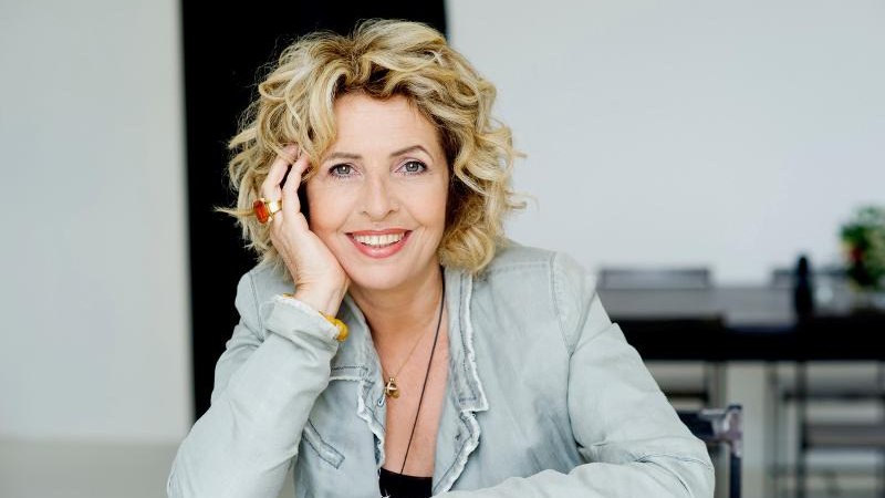 Frau mit lockigem Haar lächelt, stützt den Kopf auf die Hand. Sie trägt eine helle Jacke und sitzt an einem Tisch., © Backnang Frau mit lockigem Haar lächelt, stützt den Kopf auf die Hand. Sie trägt eine helle Jacke und sitzt an einem Tisch., © Backnang