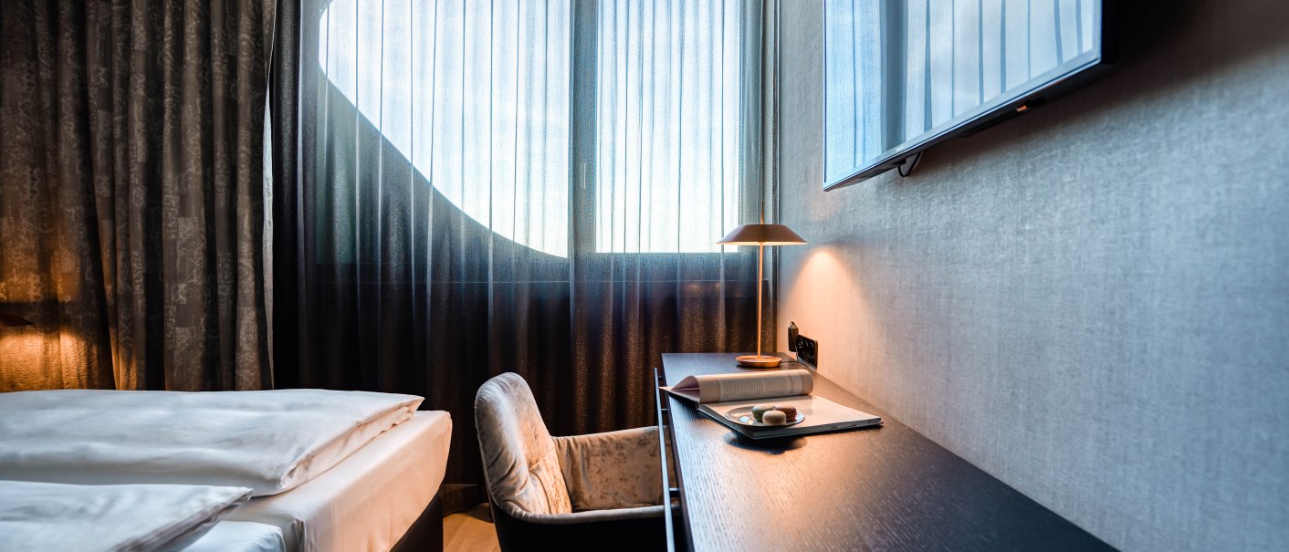 Modernes Hotelzimmer mit elegantem Schreibtisch, gemütlichem Stuhl, Doppelbett und großem Fenster mit Vorhängen. Ein Fernseher hängt an der Wand., © CLOUD N°7 APARTMENTS GmbH