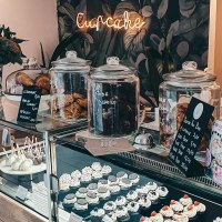 Ein Café mit einer Glasvitrine voller Cupcakes und Kekse. Ein Neon-Schild mit 'Cupcake' leuchtet im Hintergrund. Blumen schmücken den Tresen., © Cupcakes & Bagels, Stuttgart