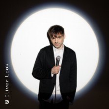 Ein Mann in einem schwarzen Anzug h&auml;lt ein Mikrofon und steht vor einem hellen Lichtkegel, der ihn beleuchtet., &copy; links im Bild