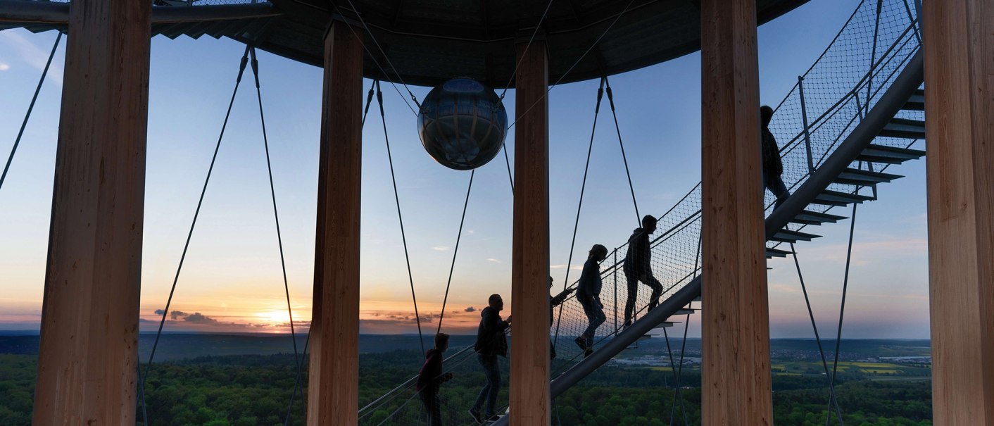Menschen steigen eine Wendeltreppe im Schönbuchturm hinauf, umgeben von Holzsäulen. Im Hintergrund ist ein Sonnenuntergang über einer weiten Landschaft zu sehen., © Stuttgart-Marketing GmbH, Martina Denker