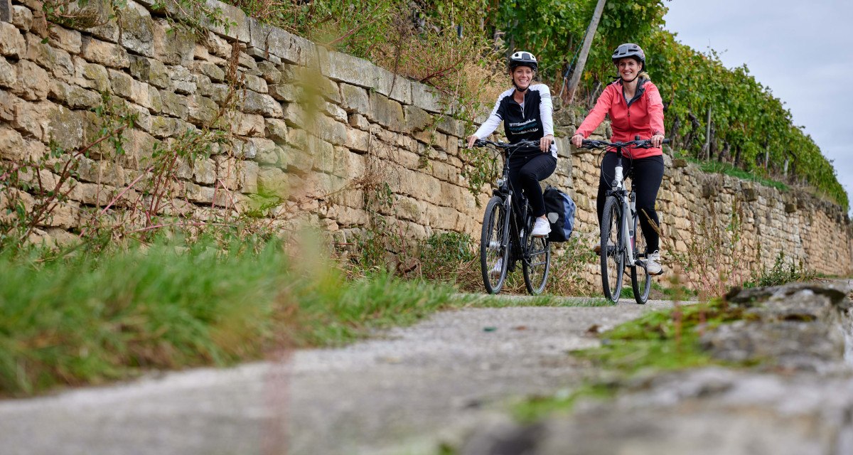 Zwei Frauen radeln lächelnd auf einem Weg neben einer alten Steinmauer, umgeben von grüner Vegetation., © Kraichgau Stromberg Tourismus e.V.-Christian Ernst Zwei Frauen radeln lächelnd auf einem Weg neben einer alten Steinmauer, umgeben von grüner Vegetation., © Kraichgau Stromberg Tourismus e.V.-Christian Ernst