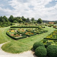 Ein weitläufiger Garten mit symmetrischen Blumenbeeten und gepflegten Hecken, umgeben von Bäumen und einem blauen Himmel., © HfWU, Manuel stark