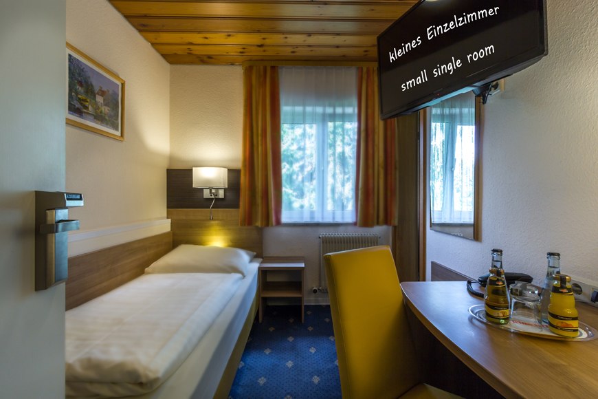 Kleines Einzelzimmer mit Bett, Tisch, Stuhl und Fernseher. Helle Wände, Holzmöbel und ein Fenster mit Vorhängen., © Römerhof Hotelbetriebs GmbH